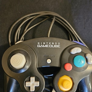May include: A black Nintendo GameCube video game controller featuring a white analog stick, a gray D-pad, a yellow C-stick, a large green A button, a red B button, gray X and Y buttons, a white Start/Pause button, and a black wired connection cable.