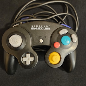 May include: A black Nintendo GameCube controller featuring a white analog stick, a yellow C-stick, a large green A button, a red B button, gray X and Y buttons, a gray D-pad, a white Start/Pause button, and a black wired cable.