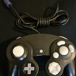 May include: A black video game controller featuring a white analog stick, a white D-pad, and white action buttons. The controller includes a black wired cable with a purple connector plug at the end, designed for use with a gaming console system.