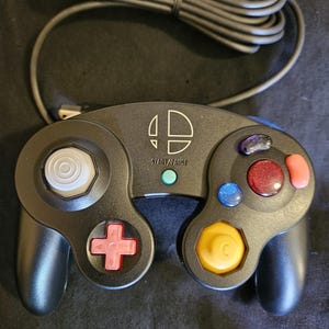 May include: A black wired game controller featuring a white Super Smash Bros logo and the text START/PAUSE above a teal button. It includes a white analog stick, a yellow C-stick, a red D-pad, and four colored action buttons in blue, red, and pink.