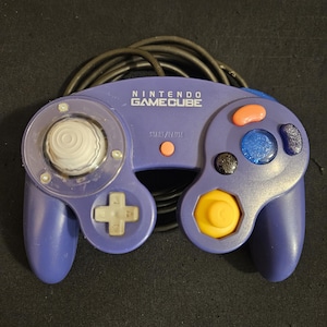 May include: A purple Nintendo GameCube video game controller featuring a white analog stick, a yellow C-stick, a gray D-pad, a red Start/Pause button, and various action buttons including a large blue button, all connected to a black cord.