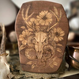 May include: A wooden plaque featuring a deer skull with antlers, surrounded by flowers and a snake. The artwork is intricately burned into the wood, creating a detailed, rustic design. The color palette is primarily brown and beige.