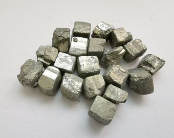 May include: A collection of small, cubic pyrite crystals. The crystals are a silvery-grey color and have a metallic sheen. They are arranged in a loose pile on a white background.