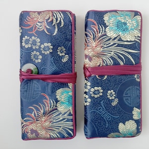 May include: Two navy blue fabric cases with floral embroidery in teal, gold, and red. Each case is wrapped with a claret ribbon. One case has a jade-coloured button closure. The cases are likely for storing items.