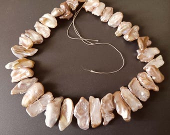 Könnte beinhalten: Eine Kette aus unregelmäßig geformten Perlen in Creme-, Braun- und Goldtönen. Die Perlen sind auf einem dünnen, cremefarbenen Faden aufgefädelt. Der Hintergrund ist dunkel und einfarbig. Die Perlen sind etwa 1-2 cm lang.