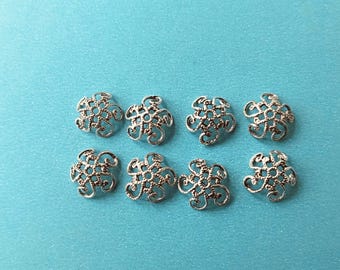 Könnte beinhalten: Acht silberfarbene Filigran-Perlenkappen auf einer türkisfarbenen Oberfläche angeordnet. Jede Kappe hat ein florales Design mit komplizierten Details. Diese dekorativen Kappen sind ideal für Schmuckherstellung und Bastelprojekte.