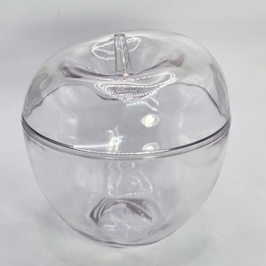 May include: A clear, apple-shaped container with a lid. The container is made of transparent plastic and has a stem-like protrusion on top. The lid fits snugly around the middle of the apple shape. The container is empty.