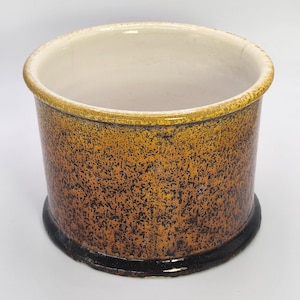 Puede incluir: Un recipiente cilíndrico de cerámica con un exterior moteado marrón y negro. El borde y el interior son de color blanco crema, y la base es de color marrón oscuro. Esta pieza decorativa podría usarse como maceta o contenedor de almacenamiento.
