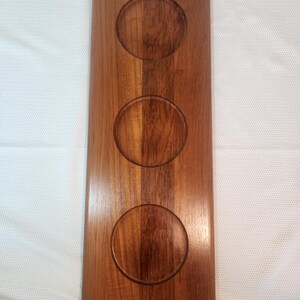 Puede incluir: Una bandeja rectangular de madera con tres hendiduras circulares. La bandeja está hecha de madera de tonos cálidos con vetas visibles. Los bordes son redondeados y la superficie es lisa y pulida.