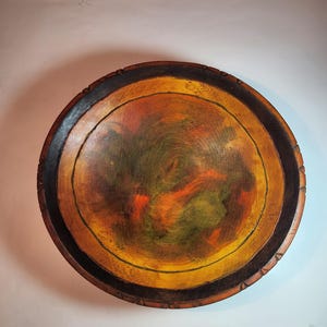 May include: A round wooden bowl with a dark brown rim and a painted interior. The interior is painted in shades of yellow, orange, and green.