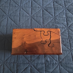 May include: A rectangular wooden box with a polished finish, featuring a two-tone brown grain pattern. The lid is constructed with a puzzle-piece design, where one section is shaped like a jigsaw piece that fits into the main body of the box.