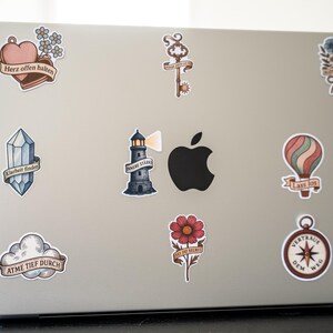 May include: A silver laptop covered in decorative stickers. Stickers include a heart with text, a key, a bouquet of flowers, a crystal, a lighthouse, a hot air balloon, a flower, a compass, and a cloud with text.