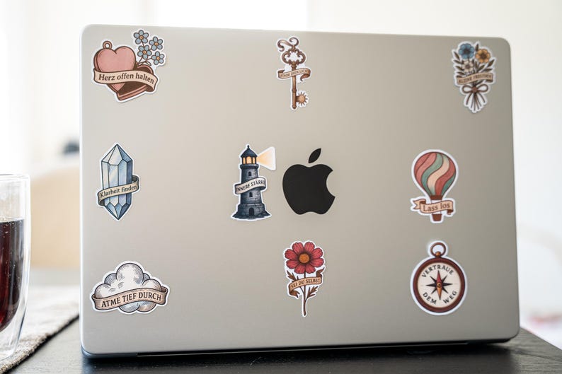 May include: A silver laptop covered in decorative stickers. Stickers include a heart with text, a key, a bouquet of flowers, a crystal, a lighthouse, a hot air balloon, a flower, a compass, and a cloud with text.