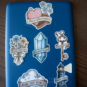 May include: A collection of decorative stickers on a blue surface. The stickers feature various designs, including a heart with the text "Herz offen halten", a bouquet of flowers, a crystal, a key, a cloud, and a lighthouse, each with German text.