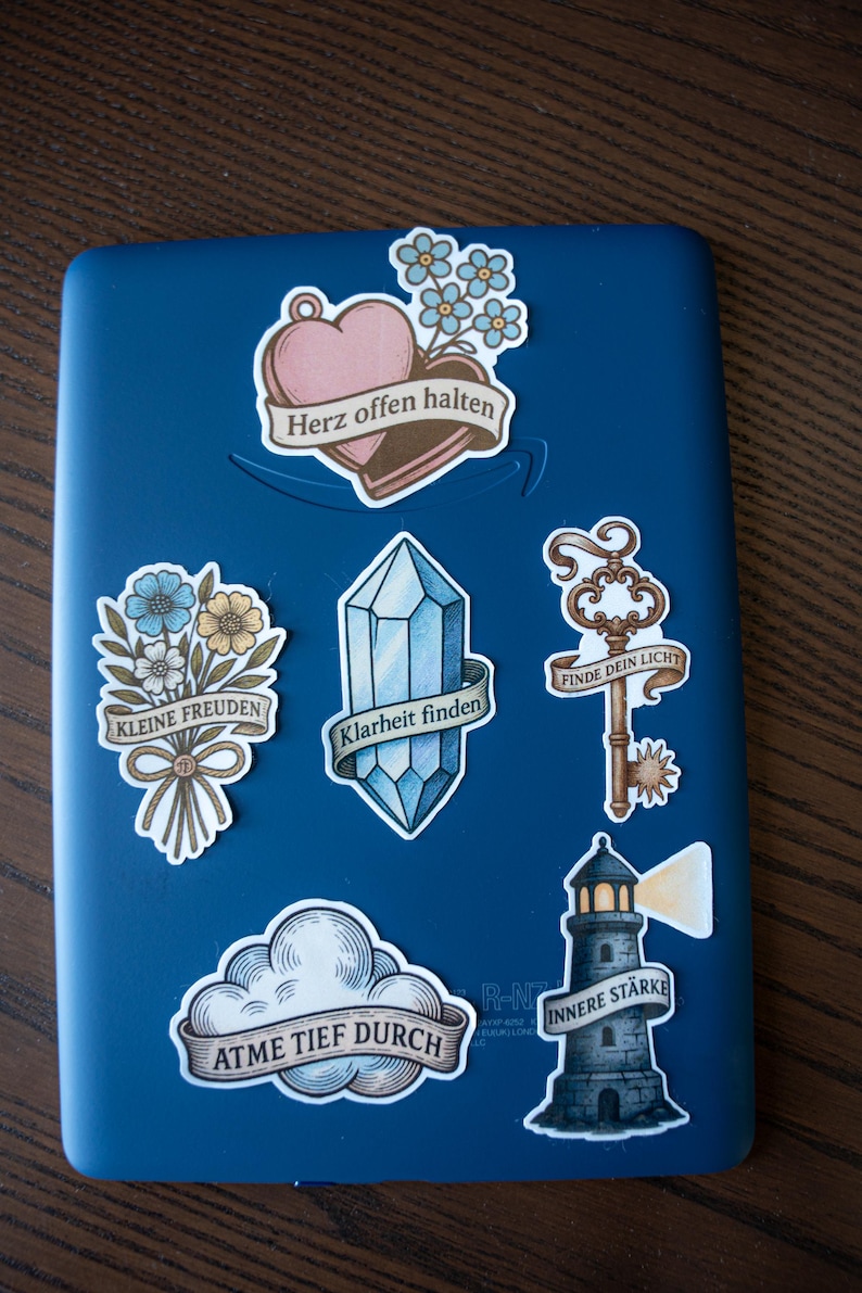 May include: A collection of decorative stickers on a blue surface. The stickers feature various designs, including a heart with the text "Herz offen halten", a bouquet of flowers, a crystal, a key, a cloud, and a lighthouse, each with German text.
