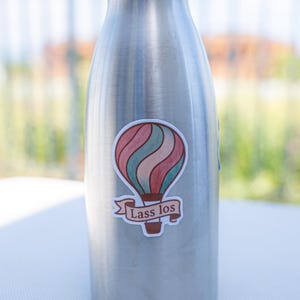 May include: A stainless steel water bottle with a black cap. A sticker of a hot air balloon in red, teal, and pink with the text "Lass los" is affixed to the bottle. The bottle is on a white surface.