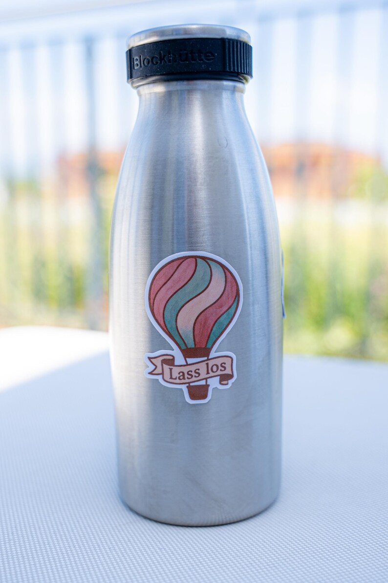 May include: A stainless steel water bottle with a black cap. A sticker of a hot air balloon in red, teal, and pink with the text "Lass los" is affixed to the bottle. The bottle is on a white surface.