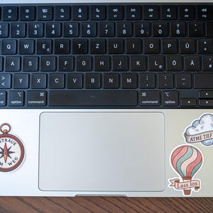May include: A silver laptop with a black keyboard and several stickers. Stickers include a red flower with text, a compass with text, a cloud with text, and a hot air balloon with text.