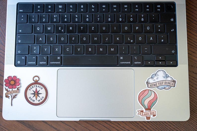 May include: A silver laptop with a black keyboard and several stickers. Stickers include a red flower with text, a compass with text, a cloud with text, and a hot air balloon with text.