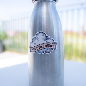 May include: A stainless steel water bottle with a black cap. A sticker with a cloud design and the text "ATME TIEF DURCH" is affixed to the bottle. The bottle is cylindrical and appears to be empty.