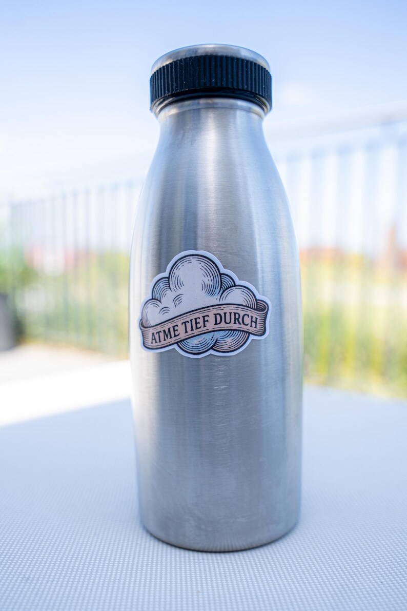 May include: A stainless steel water bottle with a black cap. A sticker with a cloud design and the text "ATME TIEF DURCH" is affixed to the bottle. The bottle is cylindrical and appears to be empty.