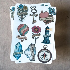 May include: A stack of illustrated stickers featuring various designs. These include a hot air balloon, a lighthouse, a compass, a key, a heart, and a crystal. Each design has a banner with German text. The stickers are on a neutral background.
