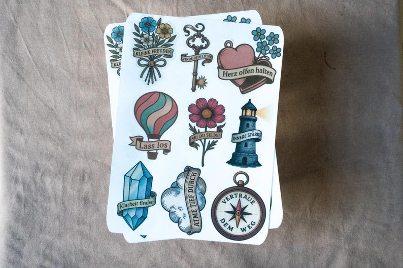 May include: A stack of illustrated stickers featuring various designs. These include a hot air balloon, a lighthouse, a compass, a key, a heart, and a crystal. Each design has a banner with German text. The stickers are on a neutral background.