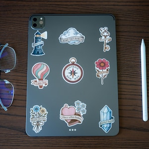 May include: A gray tablet covered in various stickers, including a lighthouse, hot air balloon, compass, and flower. A pair of glasses and a white stylus are placed next to the tablet on a wooden surface.