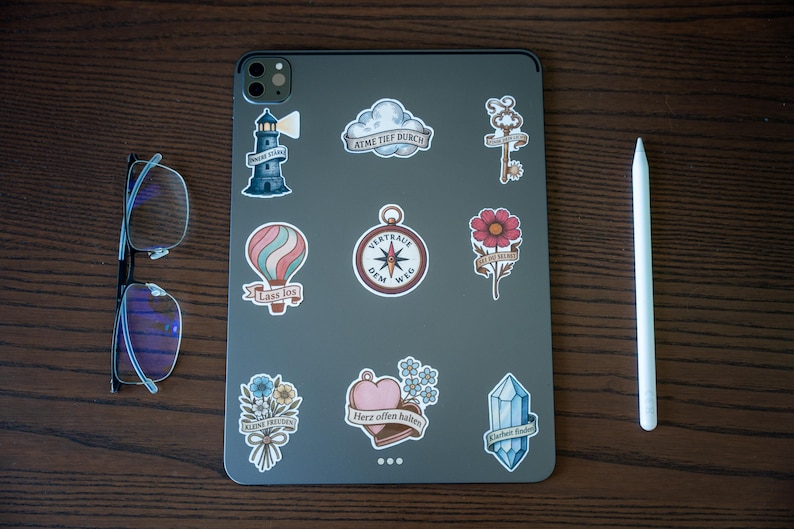 May include: A gray tablet covered in various stickers, including a lighthouse, hot air balloon, compass, and flower. A pair of glasses and a white stylus are placed next to the tablet on a wooden surface.