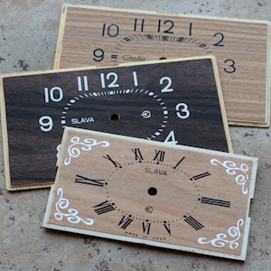 May include: Three vintage clock faces with wooden textures. One is light brown with black numerals, another is dark brown with white numerals, and the third is light brown with Roman numerals. The word "SLAVA" is visible on two faces.