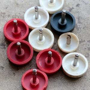 Könnte beinhalten: Neun runde, weiße, rote und schwarze Kunststoffknöpfe mit einem Metallpin in der Mitte. Die Knöpfe sind in einem dreieckigen Muster angeordnet.