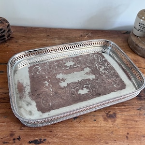 May include: A rectangular silver-plated serving tray with a decorative openwork gallery rim. The centre of the tray features an intricate, tarnished copper-toned engraved pattern with scrollwork details and a worn, aged metallic finish.