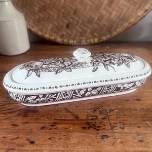 May include: An oval ceramic butter dish with a cream-coloured base and a matching lid. The dish features a dark brown transferware pattern depicting fruit and foliage, with a small knob handle on the centre of the lid and a decorative border.