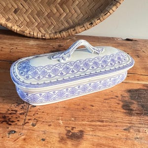 May include: A white and blue ceramic trinket box with a lid and handle. The box is rectangular with rounded edges and a blue floral pattern. It rests on a wooden surface. The box is approximately 20 cm long.