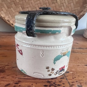 May include: A cream-colored stoneware crock with a matching lid secured by a black metal clamp. The jar features a teal band near the top, a stamped number 8, and faded red and green floral-style painted accents on the exterior surface.