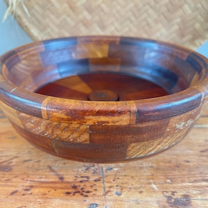 May include: A round wooden bowl with a mosaic pattern of various wood tones. The bowl has a wide rim and a dark interior. The bowl is sitting on a wooden surface.