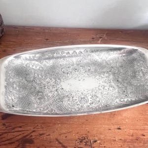 May include: A rectangular silver-toned metal tray featuring an intricate, embossed floral and scrollwork pattern across its surface. The tray has smooth, slightly raised edges and a polished finish, with a central blank area surrounded by detailed engravings.