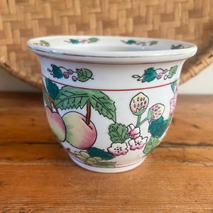 May include: White ceramic planter with a colorful fruit and floral design. The planter features a red rim and a detailed pattern of apples, strawberries, and flowers. The background includes a woven basket and a wooden surface.