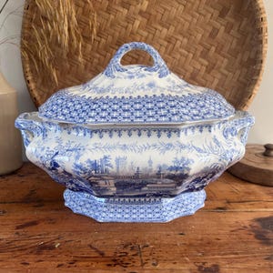 May include: A white ceramic tureen with a blue transferware pattern featuring detailed landscape scenes, architectural elements, and intricate geometric borders. The lidded serving dish has two side handles and a central arched handle on the lid.