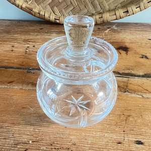 May include: A clear, cut-glass jar with a lid and a starburst design on the base. The jar has a rounded shape and sits on a wooden surface. The lid has a small handle.