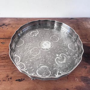 May include: A silver-colored, round serving tray with a scalloped edge and intricate floral and geometric patterns. The tray features a decorative border and a central monogram. The tray is displayed on a wooden surface.