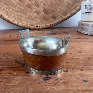 May include: An oval-shaped bowl featuring a polished silver-tone metal rim and two matching handles, set against a warm-toned wooden body with a visible grain pattern. The base is reinforced with a silver-tone metal band, creating a sturdy structure.