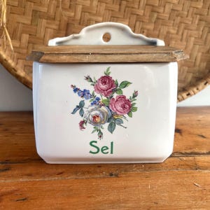 May include: A white ceramic wall-mounted salt box with a wooden lid. The front features a colorful floral bouquet of pink, blue, and white roses with green leaves. The word Sel is printed in green lettering below the floral design.