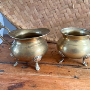 May include: Two antique brass cream jugs or small pots. One has a handle and a spout, the other is a small bowl. Both have three decorative feet and are on a wooden surface. The background includes a woven basket.