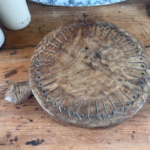 May include: A circular brown wooden board featuring a carved border of radiating petal-like shapes with small circular indentations. A small, carved wooden handle shaped like a stylized fish head extends from one side of the round wooden board.