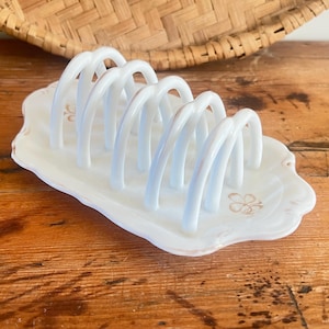 May include: A white ceramic toast rack with six arched slots, set on a scalloped base with floral accents. The rack is designed to hold toast upright. The background includes a woven basket and a wooden surface.