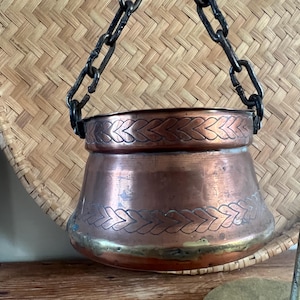 May include: A copper hanging pot with a dark metal chain. The pot has decorative etched patterns around the top and bottom. The pot is a warm copper color with a brass-colored rim.