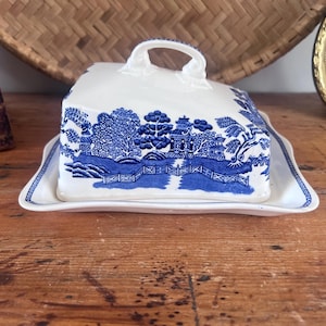 May include: A white ceramic butter dish with a blue Willow pattern design. The dish has a square base and a domed lid with a handle. The blue and white design features a landscape scene. The dish is on a wooden surface.