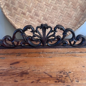 May include: A dark brown wooden architectural pediment featuring intricate scrollwork and carved floral motifs. The decorative piece has a flat base and a symmetrical design with curved flourishes extending outward from a central fan-like element.