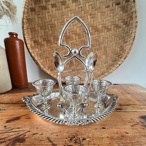 May include: A silver-plated egg cup set with four glass cups and two spoons. The set includes a decorative handle and sits on a silver tray with a scalloped edge. A woven basket and ceramic bottles are in the background.